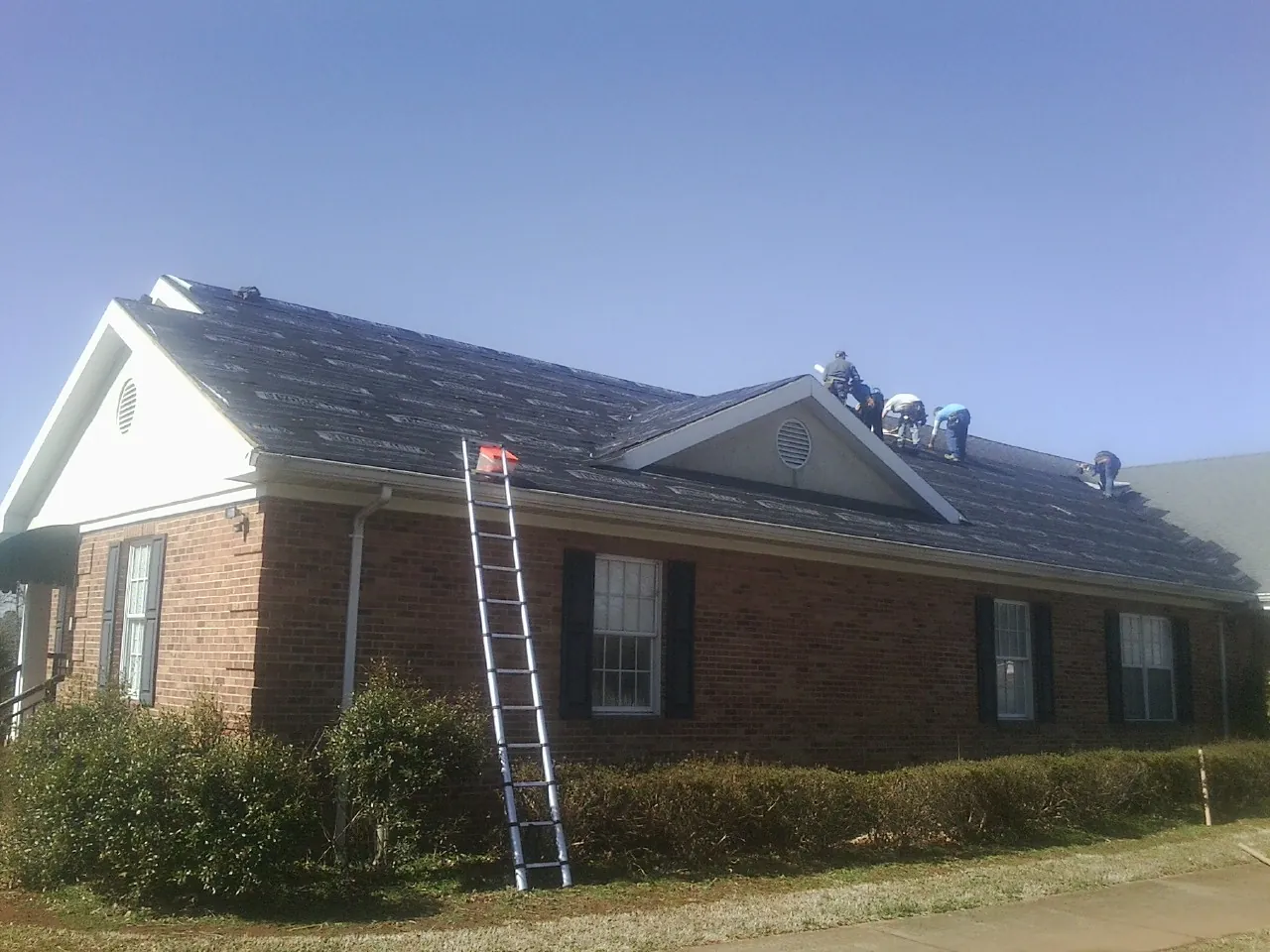 Crew tearing shingles off a brick ranch roof with a ladder set — Metal Roof Repair work by our crew in Olaton