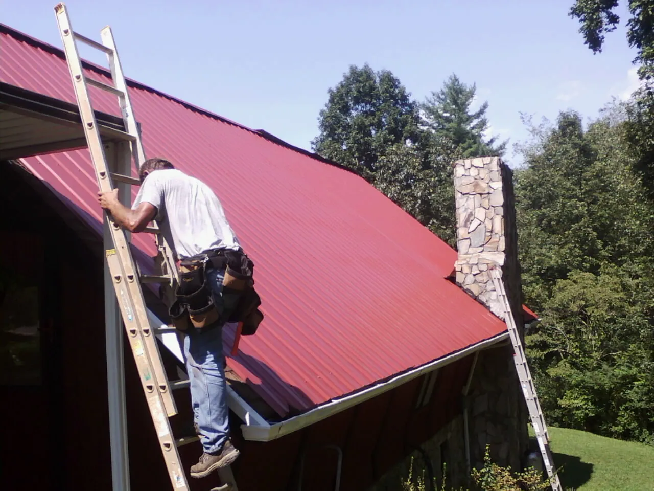 Roofer on a ladder beside a red metal roof and stone chimney — Soffit & Fascia Repair by Roofing Brothers in Olaton